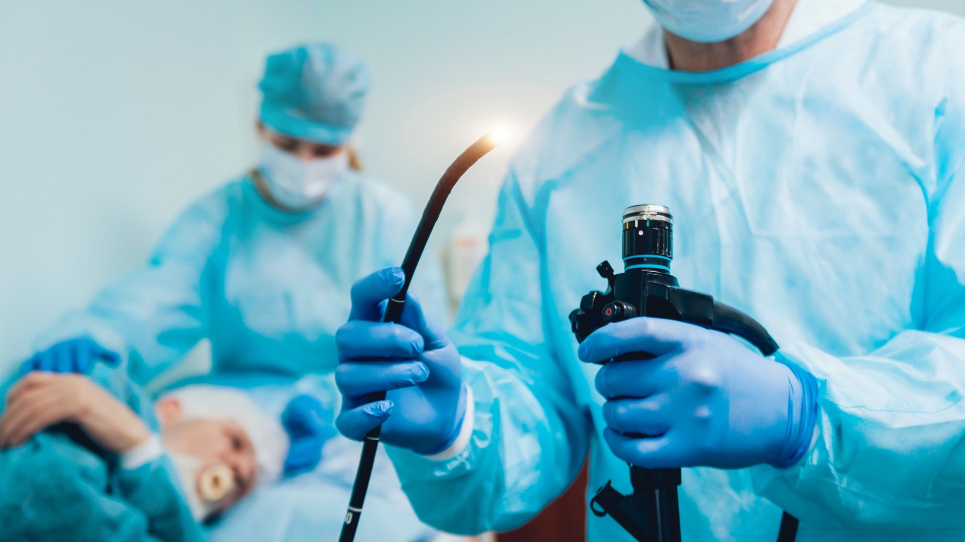 Arzt hat ein Endoskop in der Hand, im Hintergrund ist eine OTA mit einem Patienten zu sehen