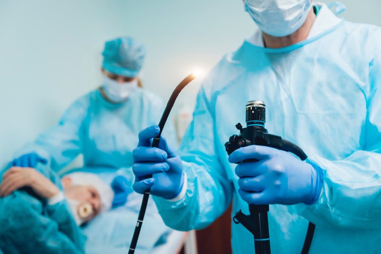 Arzt hat ein Endoskop in der Hand, im Hintergrund ist eine OTA mit einem Patienten zu sehen