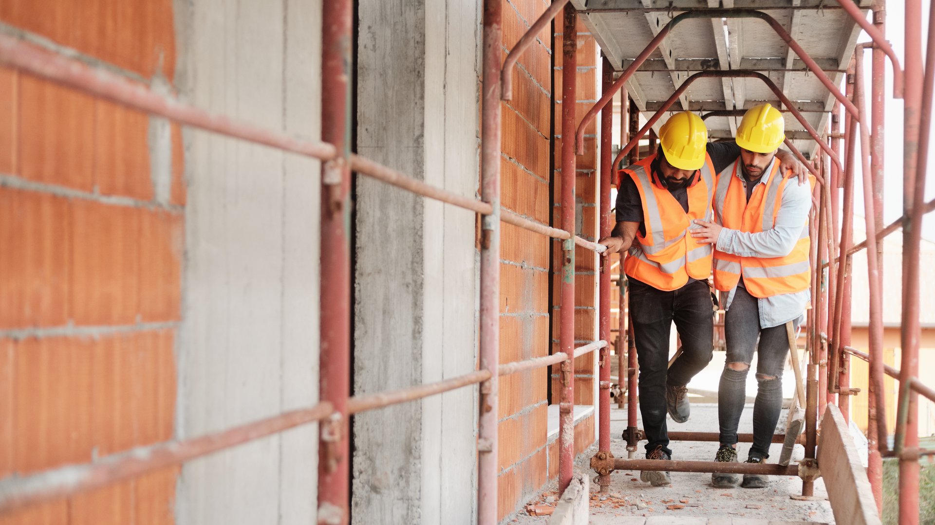 Zwei Bauarbeiter mit gelbem Schutzhelm auf einer Baustelle. Der eine stützt den Anderen, da dieser sich bei einem Berufsunfall ein Bein verletzt hat.